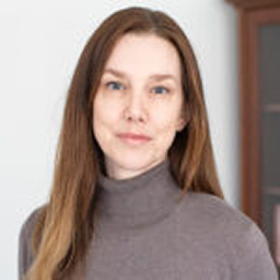 Kvinna med långt brunt hår och grå polotröja står framför en vit bakgrund.