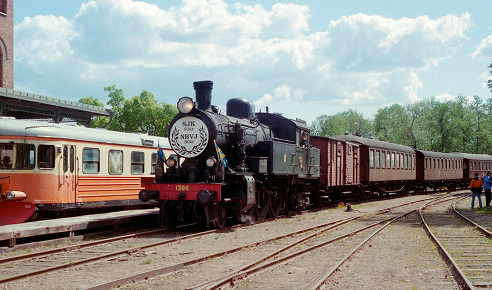 Ånglok på en järnvägsstation, omgiven av vagnar och några människor.
